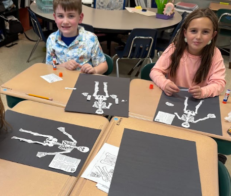 Two students sit at a table, each with a paper skeleton in front of them, working on projects.