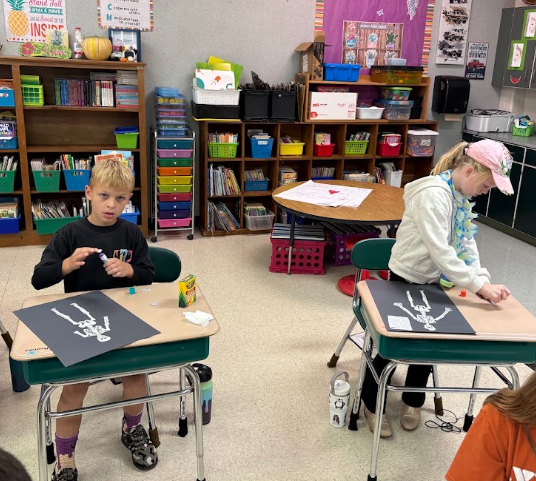 Two children sitting at desks in a classroom, one with a pencil and paper, the other with a paper skeleton.