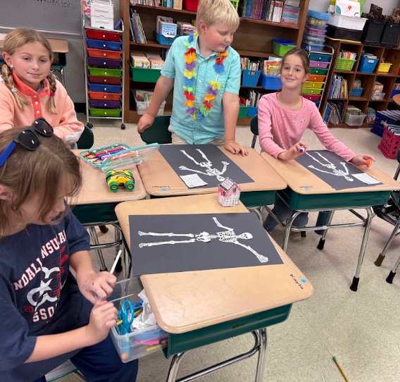 Four children seated at desks with a skeleton drawing on a paper in front of them.