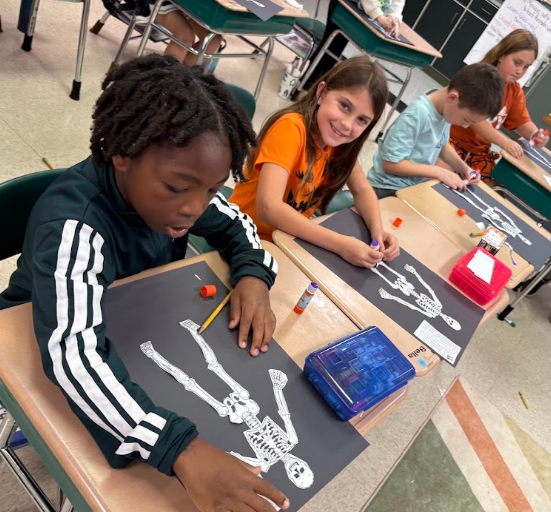Four children, seated at desks, working on skeletons drawn on black paper; one holding a pencil.