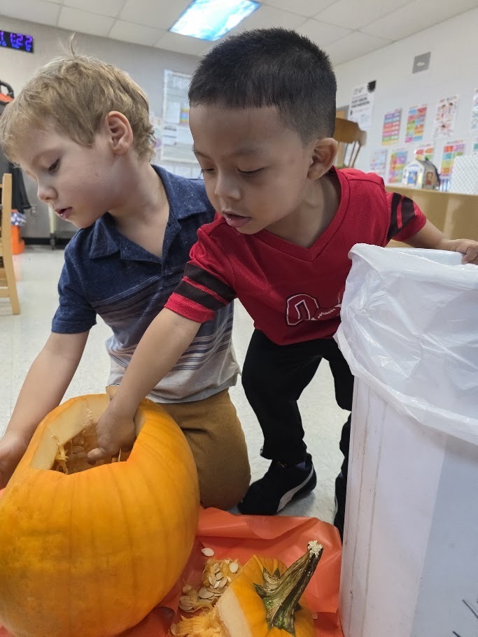 Two children scoop out pumpkin seeds. One holds a pumpkin; the other leans over a white container.