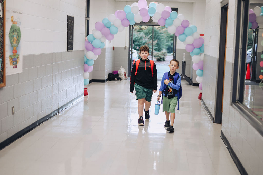 Two students walk through a hallway with a colorful balloon arch and a poster on the wall.