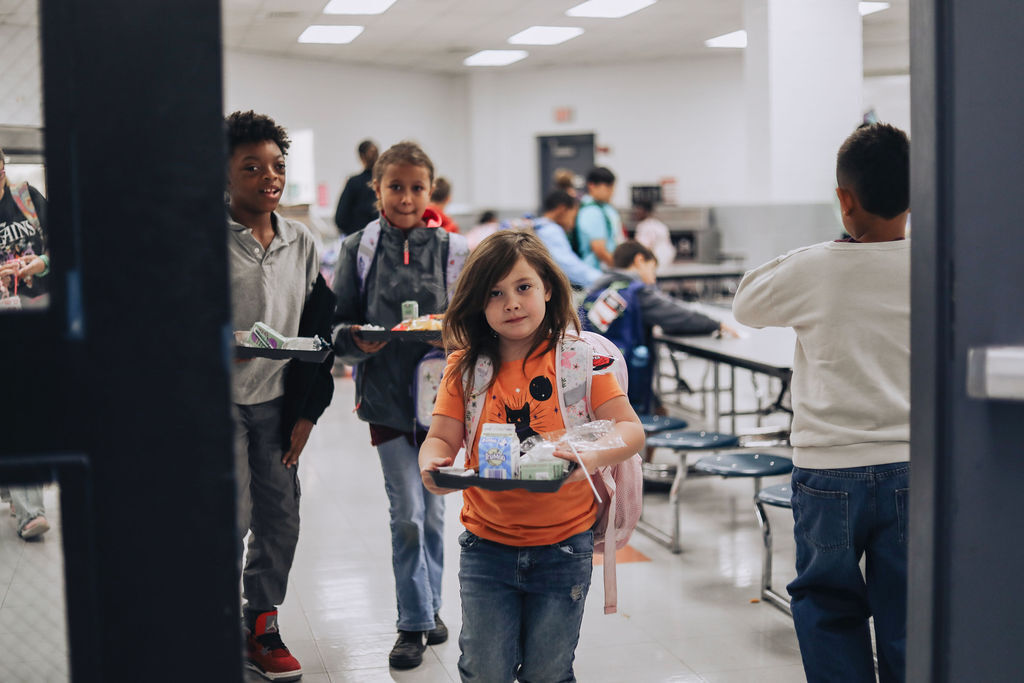 Kids with trays carrying food through a school hallway. A few more kids sit at tables in the background.