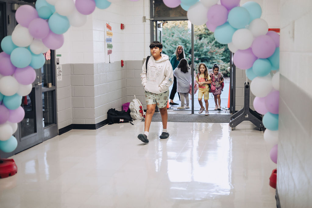 Person walking past balloons in hallway, others walking through doors, one holding hands with children.
