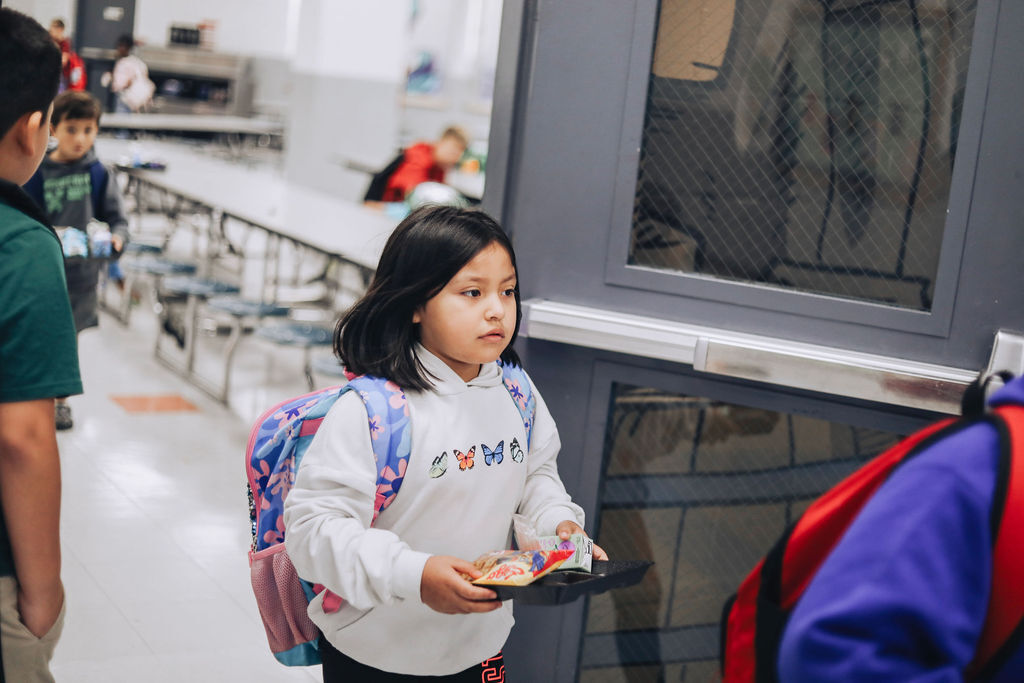 A girl with a backpack holds a book in a hallway. Behind her, a person stands at a window.