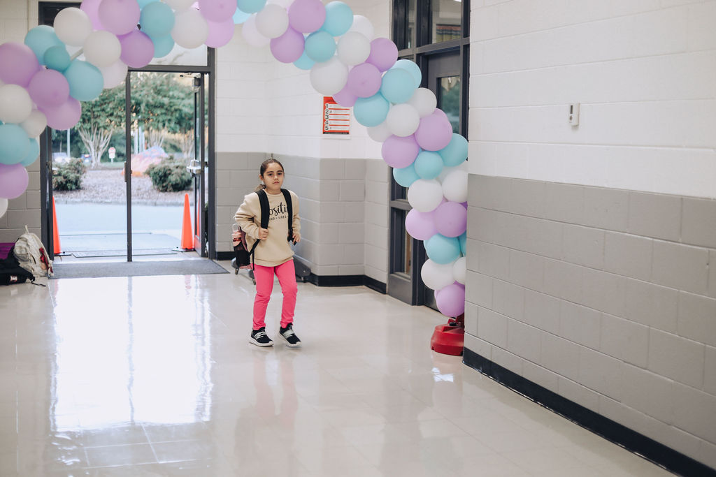 A young person wearing a backpack walks past an archway made of multicolored balloons.