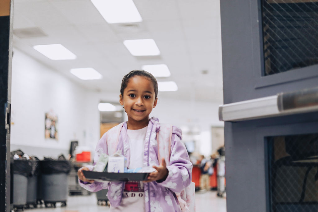 A young child holds a black tray with cups on it, smiling and standing near a door.