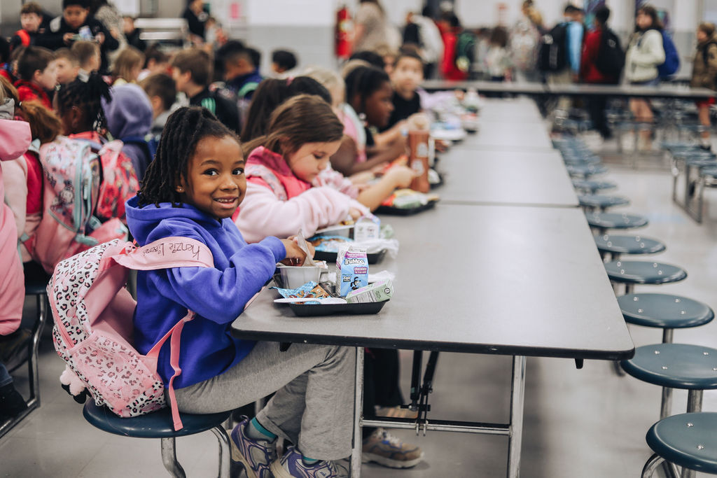 Students seated at tables with backpacks, eating lunches, in a cafeteria with chairs and tables in rows.