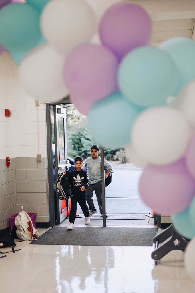 Two kids walking into a school hallway decorated with colorful balloons and a rug.