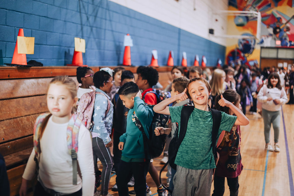 Many students in a gymnasium, some carrying backpacks, wearing winter clothes, with blue walls and orange traffic cones.