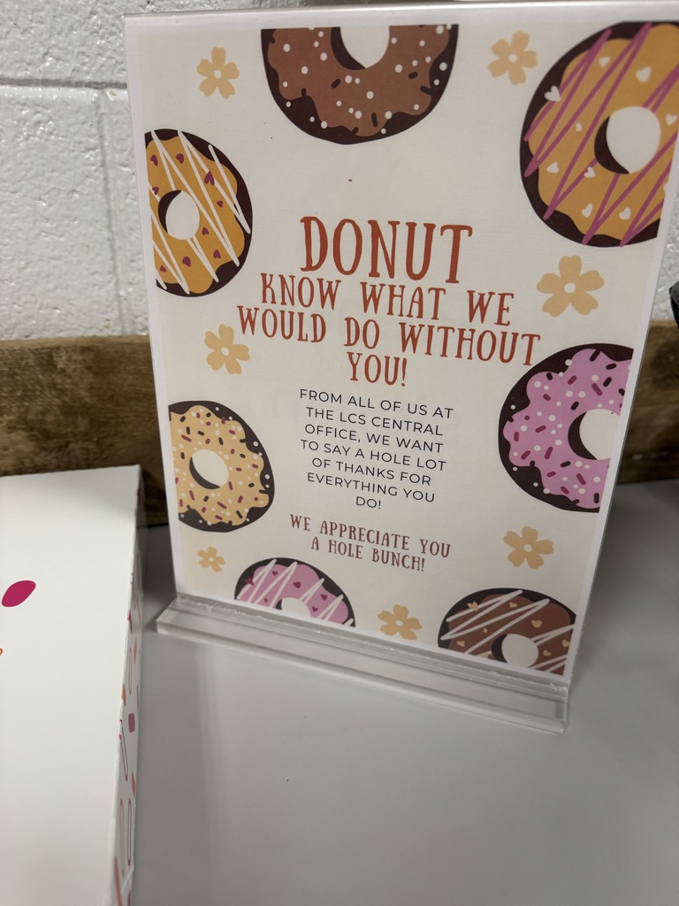 A sign displays doughnuts and reads "Donuts know what we would do without you!" A white box sits beside the sign.