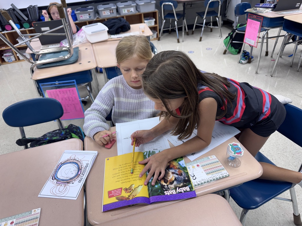 Two children in a classroom, looking at and drawing in a book, with papers and a pencil on the table.