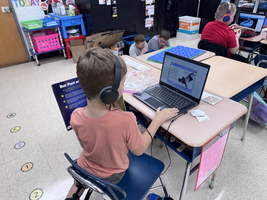 Child wearing headphones using laptop on table, other children seated at tables behind.