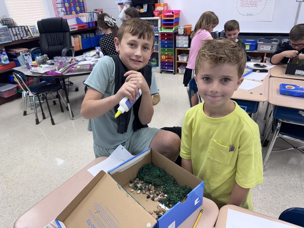 Two boys, one using a glue stick, sit by a box of craft materials on a desk.