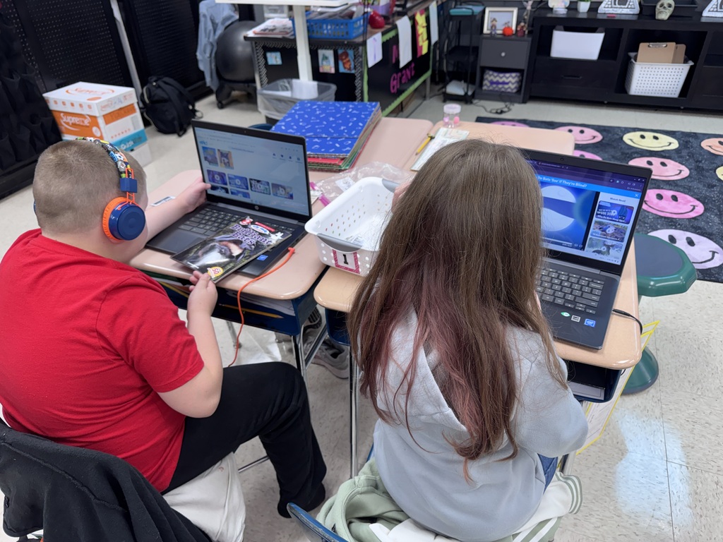Two students, one in a red shirt, one in a gray shirt, seated with laptops on desks.