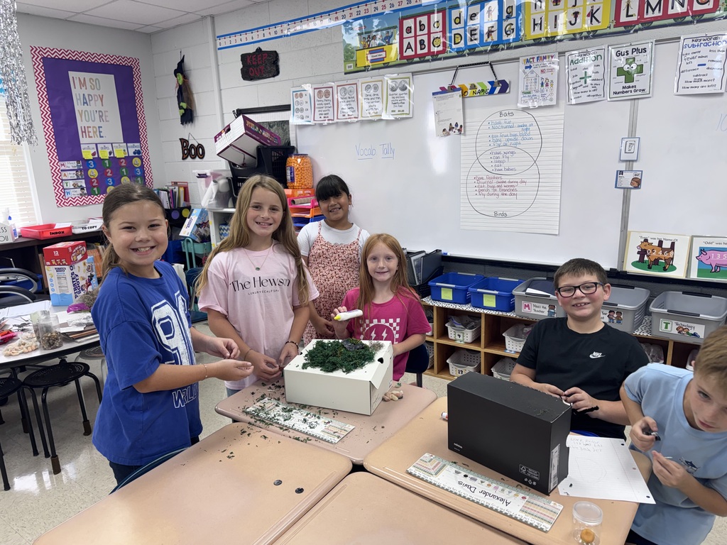 Students work together on a table with a model, plants, and a computer monitor.