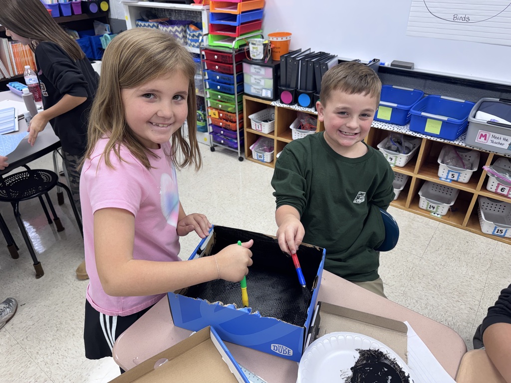 Two children in a classroom stand by a table with a blue box, holding pens and crayons.