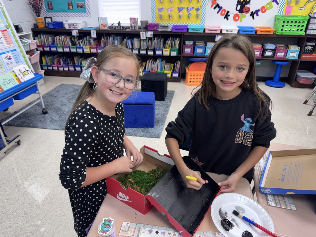 Two girls in a classroom with bookshelves, one holding a paintbrush and the other a paint palette.