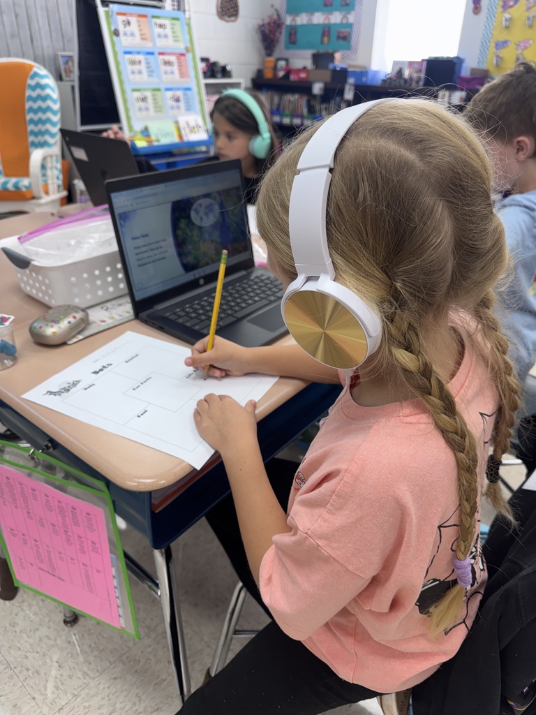 A young girl wearing headphones sits at a desk and writes on a sheet of paper.
