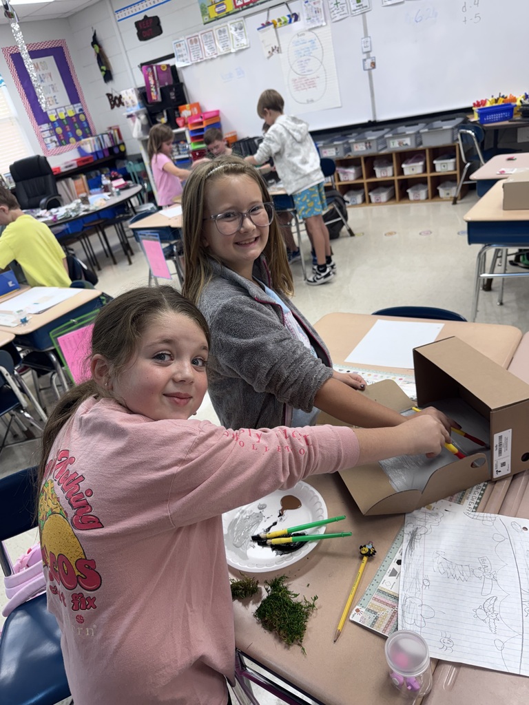 Two girls at a desk in a classroom. One holds a box with paint supplies.