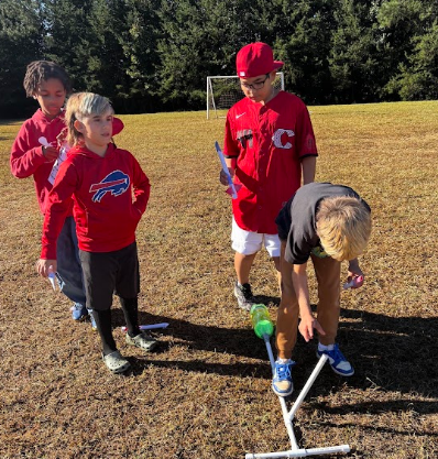 Four children outdoors, one bending to position a rocket on a stand.