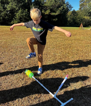 Child wearing a black shirt stomps a rocket launcher on a grassy field.