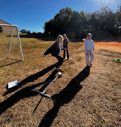 Three children play on a grassy field; one jumps on a long stick, a soccer goal is nearby.