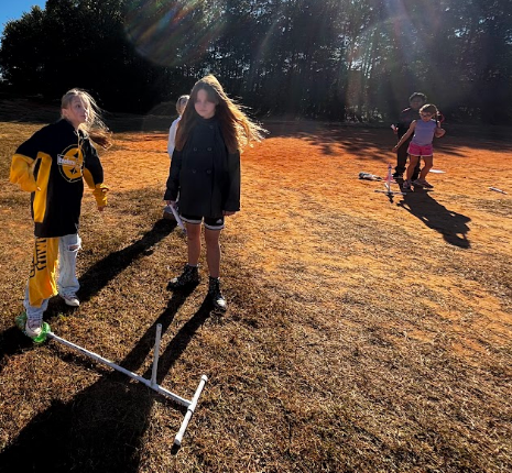Children play outside on a field with dried grass; one holds a long object. Bright sunlight and shadows.