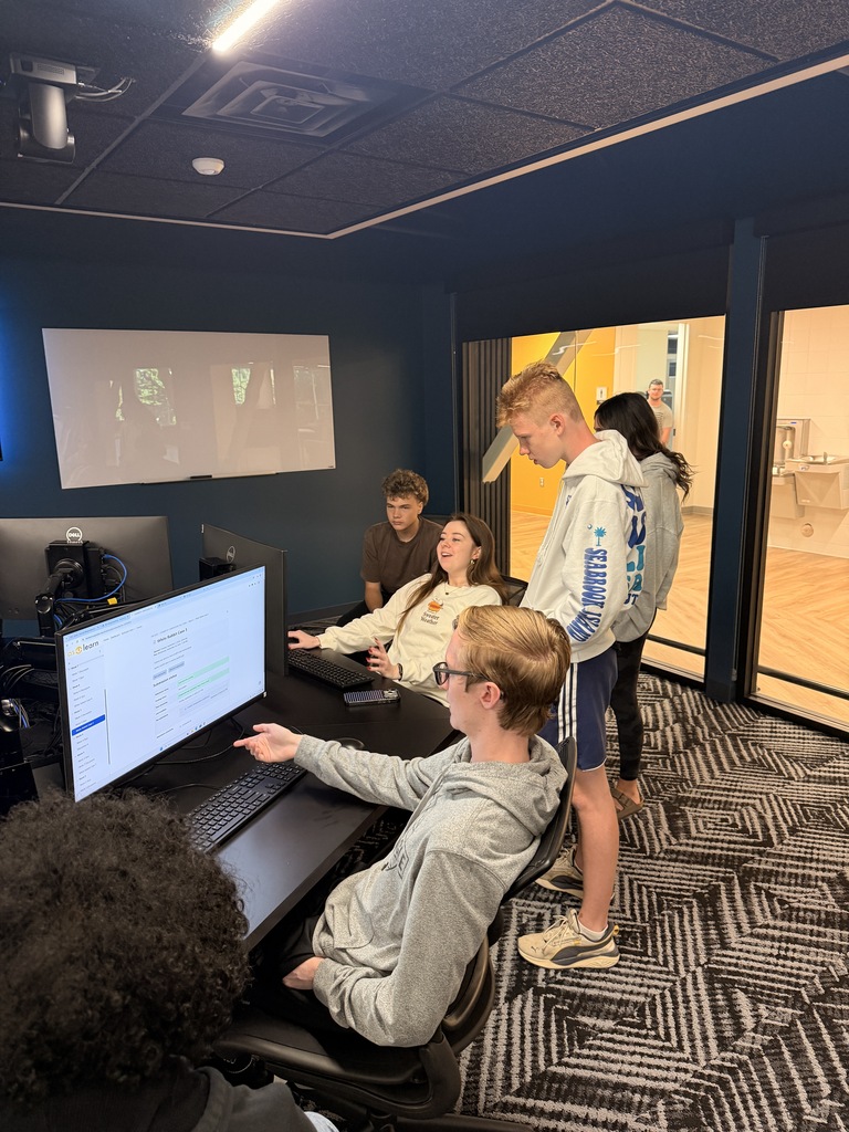 Several students sit at computers in a classroom. Two are seated; one points at a monitor.