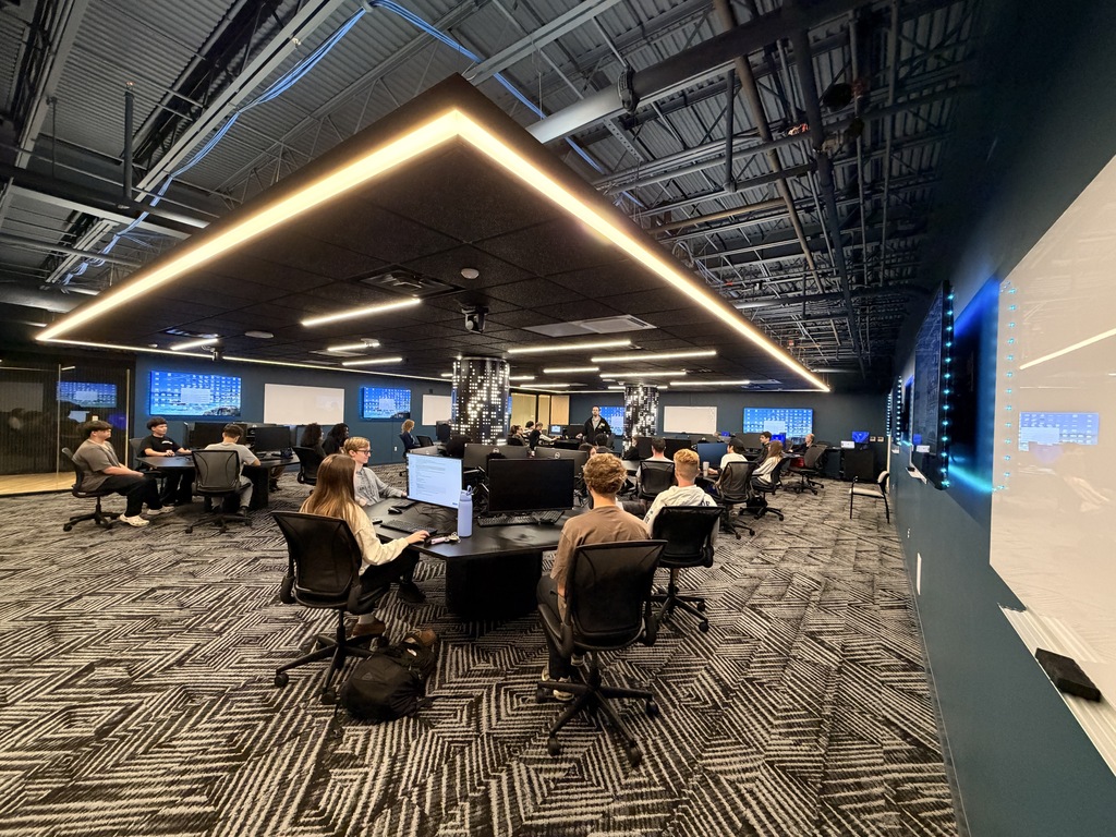 People seated around tables with monitors, in a room with carpet, wall with white board, and a ceiling with long lights.