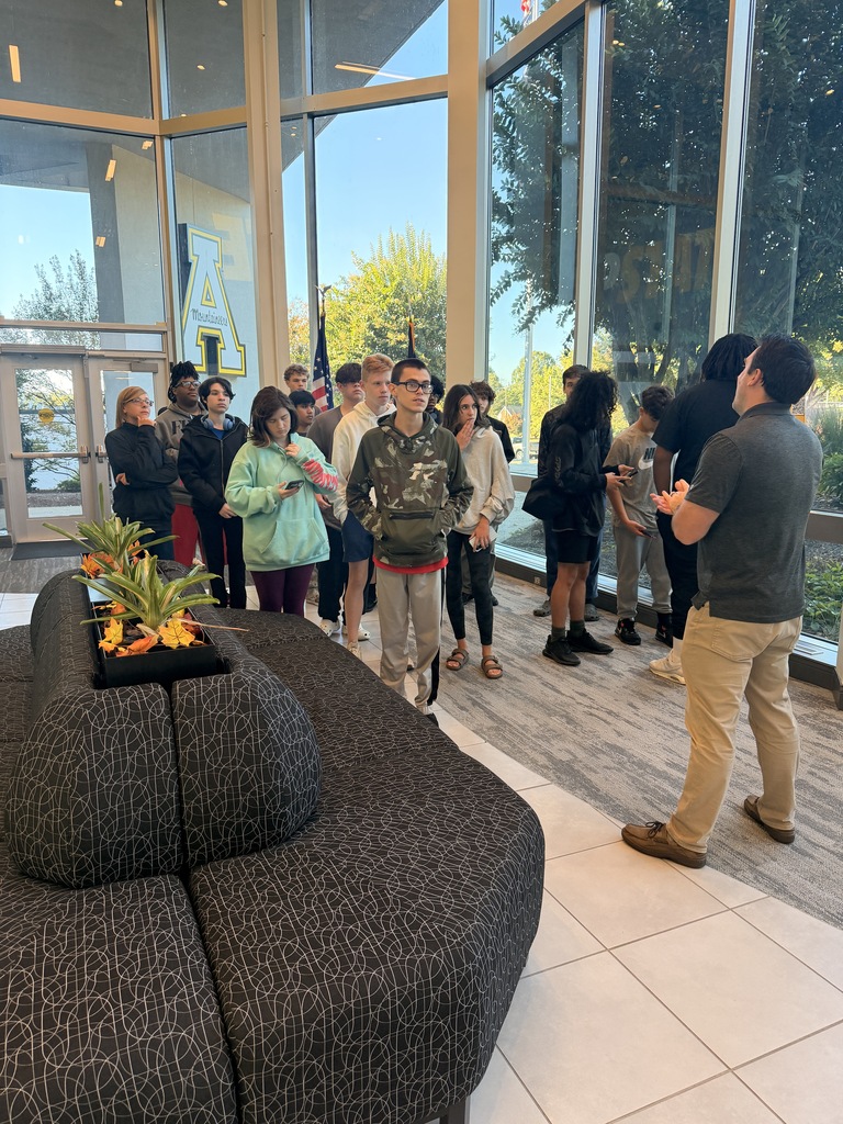 A group of people stand by a long couch near a large glass wall with trees outside.
