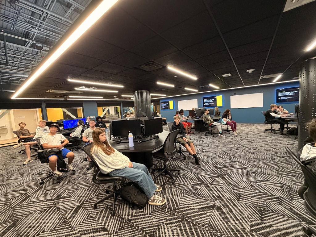 People sitting at tables in a room with black and white flooring. Monitors and chairs are visible.
