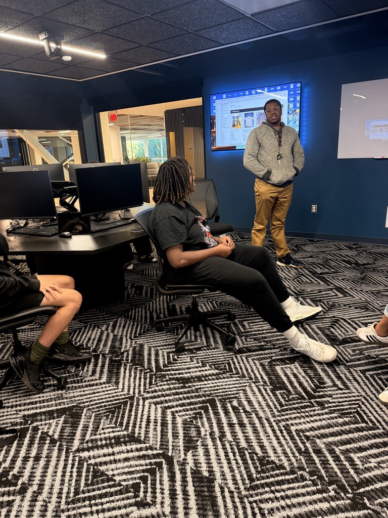 Students and an adult in a computer room, with two sitting on chairs and one standing in front of a screen.