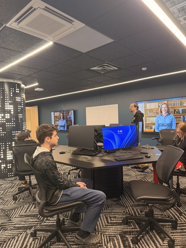 People sitting at a table with computer monitors; one person stands in front of a whiteboard.