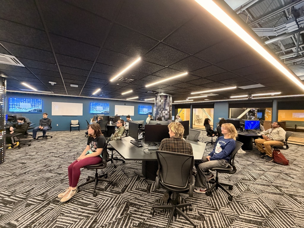 People sit around a table in a room with monitors on the wall and lights on the ceiling.