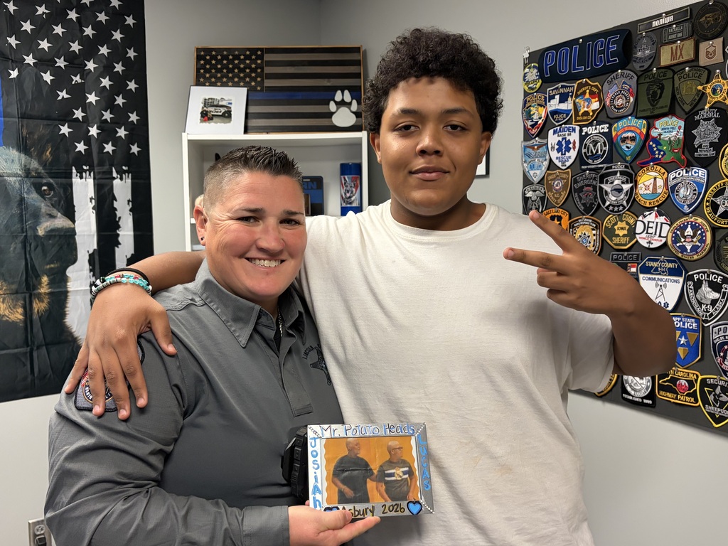 Police officer in uniform and a smiling student hold a photo album, with a wall of badges and an American flag in the background.