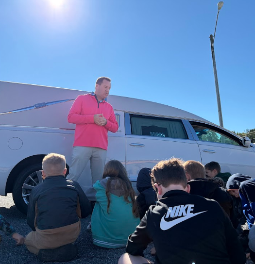Several students sitting on ground, man standing in front of white vehicle, clear sky, pole with streetlight in background.