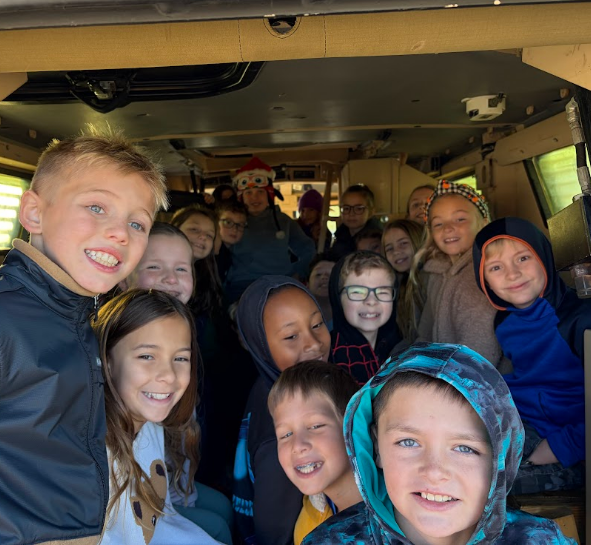 Kids in winter clothes smiling, seated inside a vehicle, some wearing hats, in front of a window.