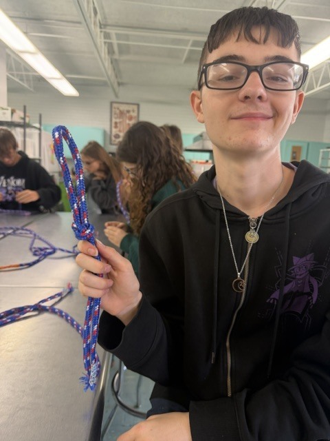 Boy holding blue rope, seated in a classroom, wearing glasses and a black sweatshirt with a purple logo.