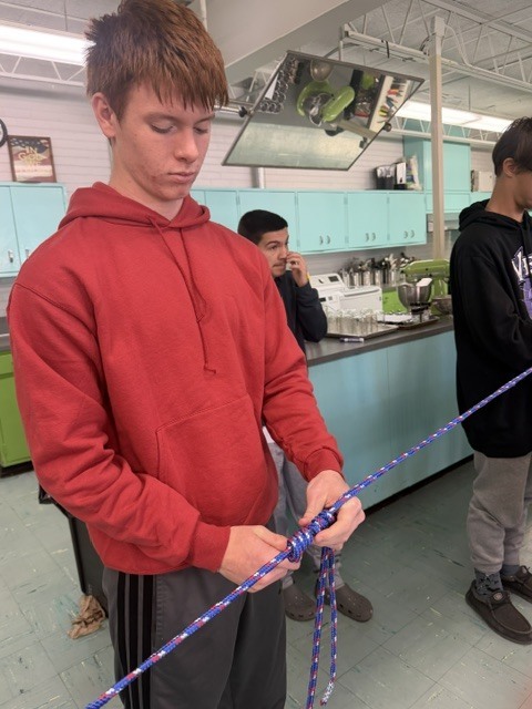 Three students in a kitchen area, one man in a red hoodie working with a rope.