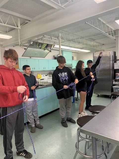 Five students tying ropes together in a lab while standing on chairs and a table.