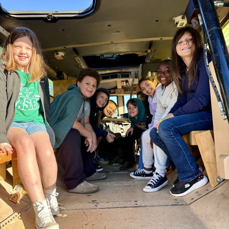 Several children seated inside a school bus, smiling at the camera, with a bench and window visible.