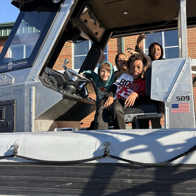 Children sit in boat, one raising arm, with boat number 509 and building in background.