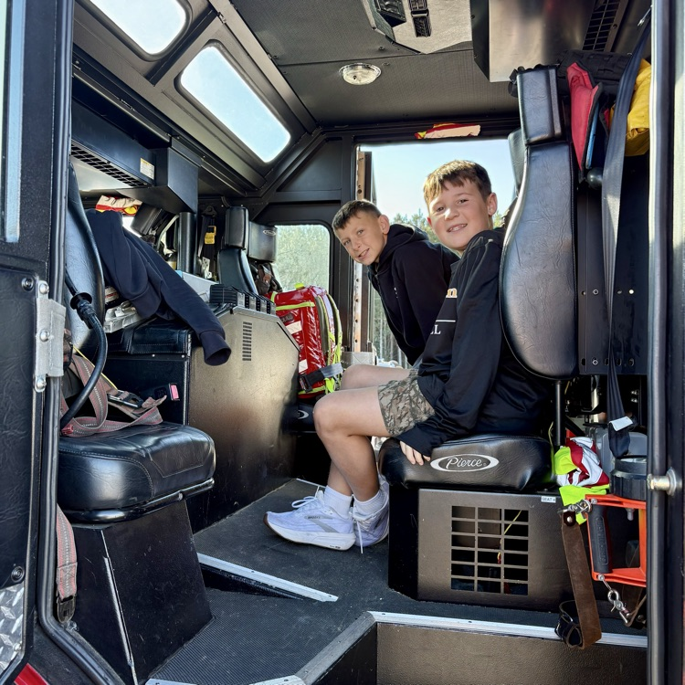 Two students sit in a firetruck's cab, one in the driver's seat, the other beside him.