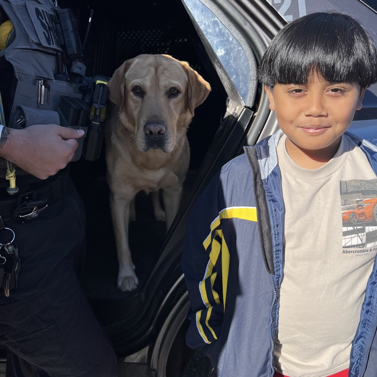 A police officer holding a leash with a standing yellow dog next to a smiling child.