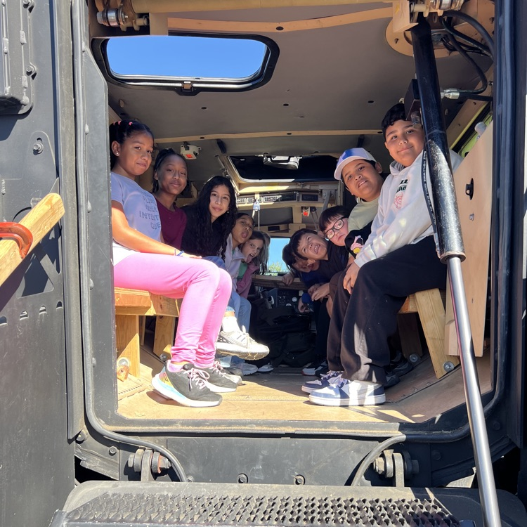 A group of students sit on benches inside a vehicle, wearing hats and sneakers, with sunlight visible through the open door.