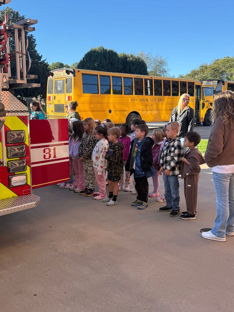 Children stand next to a red fire truck; three yellow school buses are parked in the background.
