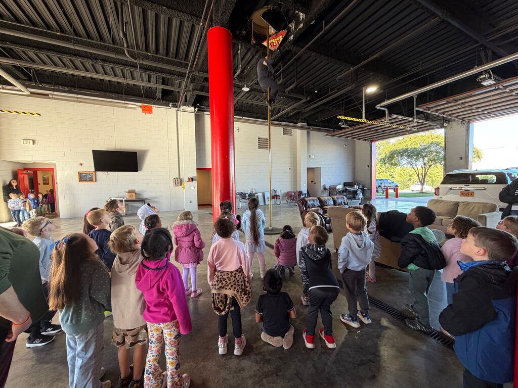 Children gathered in a fire station, some standing, some sitting, looking towards a large red pole.