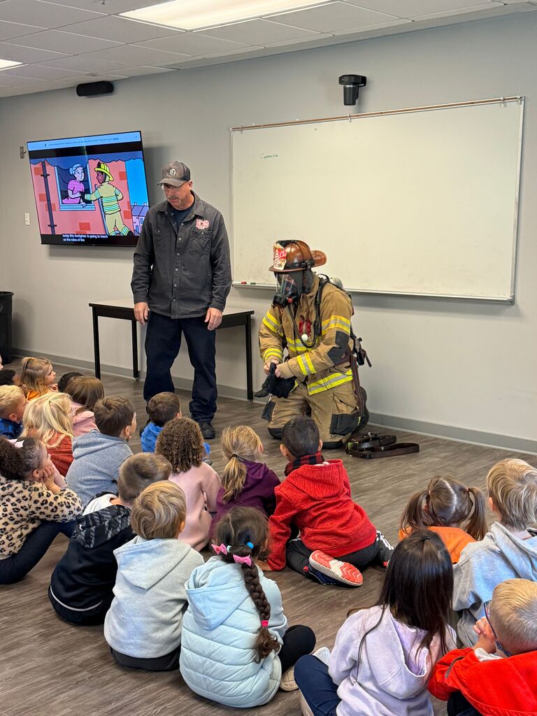 A firefighter demonstrates safety gear to a group of children sitting on the floor in a classroom.