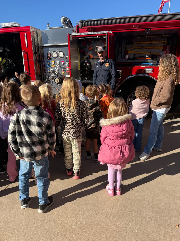 Children listen to a firefighter by a red fire truck. Some wear jackets and sneakers.